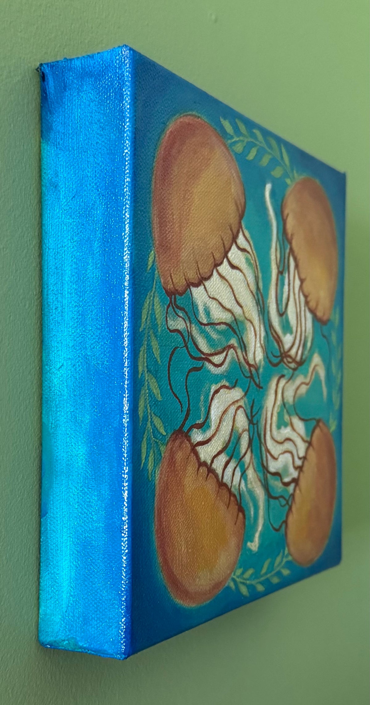 A side view of a square canvas painting of four orange jellyfish arranged symmetrically on a blue green background with green leafy accents