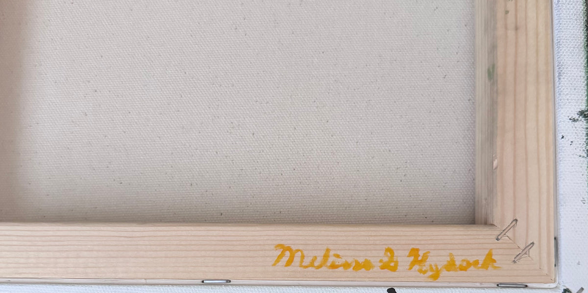 Close-up of a wooden picture frame with a yellow handwritten signature near the bottom right corner that reads, "Melissa G Hydock"