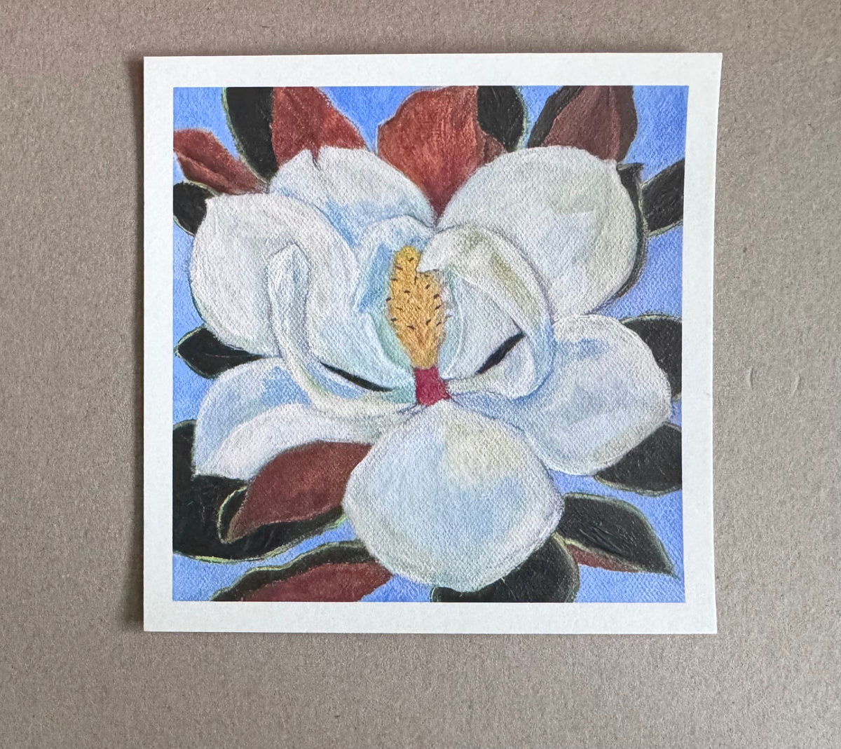 4.25in x 4.25in art print with white borders of an open magnolia flower with surrounding green and brown leaves